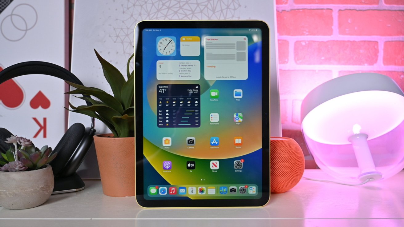 10,9-дюймовый iPad от Apple стоит на столе рядом с фиолетовой лампочкой, AirPods Max, оранжевым HomePod mini и растением.