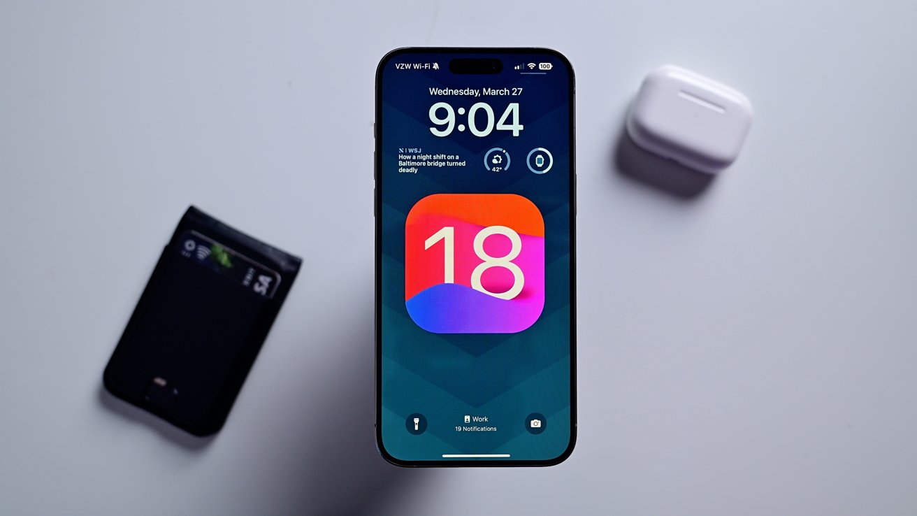 iPhone с экраном блокировки, на котором отображается значок iOS 18.  Рядом кошелек и чехол для AirPods.