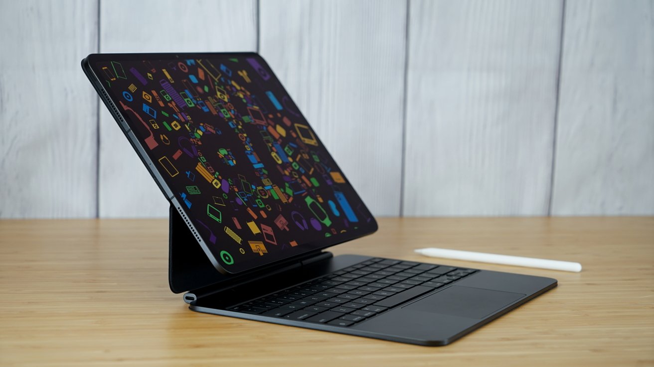 iPad Pro с цветным экраном на подставке для клавиатуры, установленной на деревянной поверхности.  Стилус лежит рядом на светлом деревянном фоне.