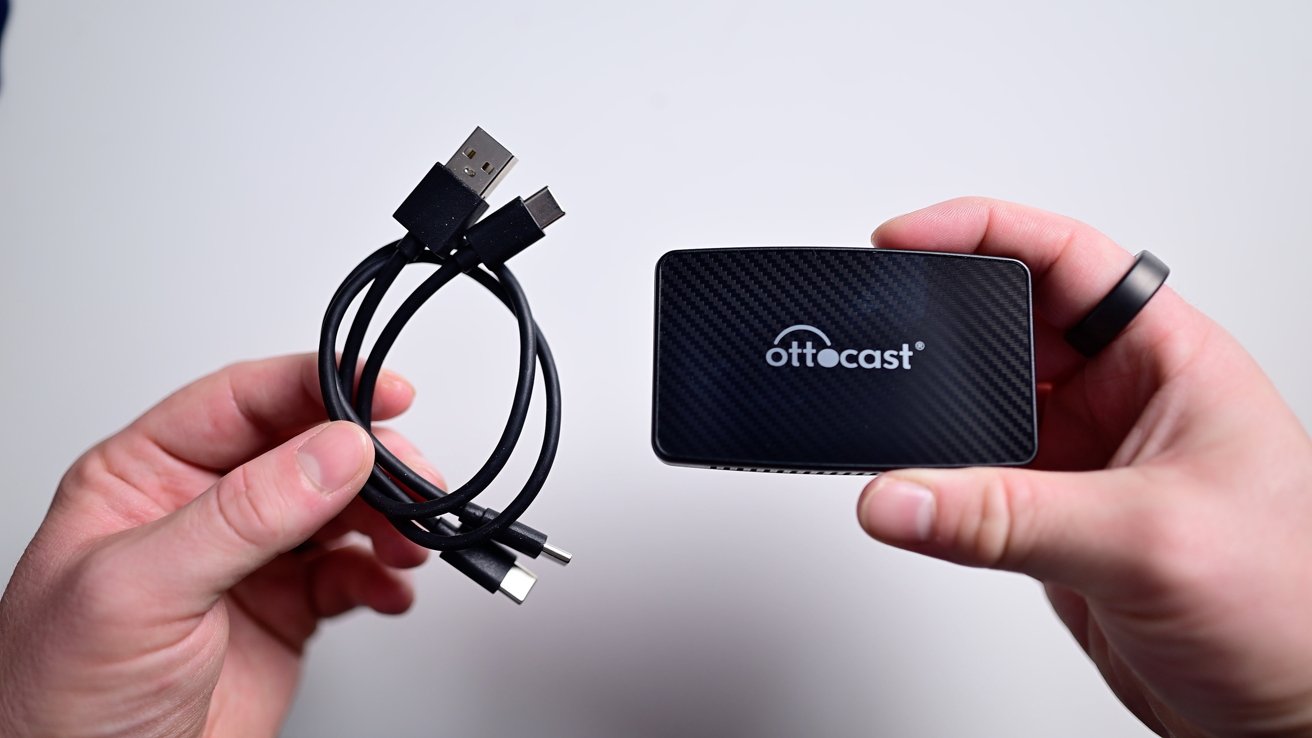 Руки держат спиральный USB-кабель и небольшое черное прямоугольное устройство с надписью «Ottocast».
