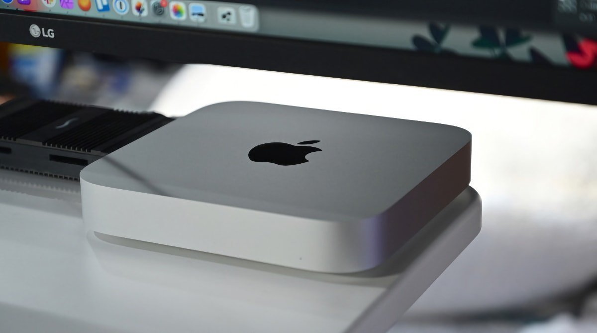 Серебристый компьютер Mac Mini под монитором LG на белом столе.