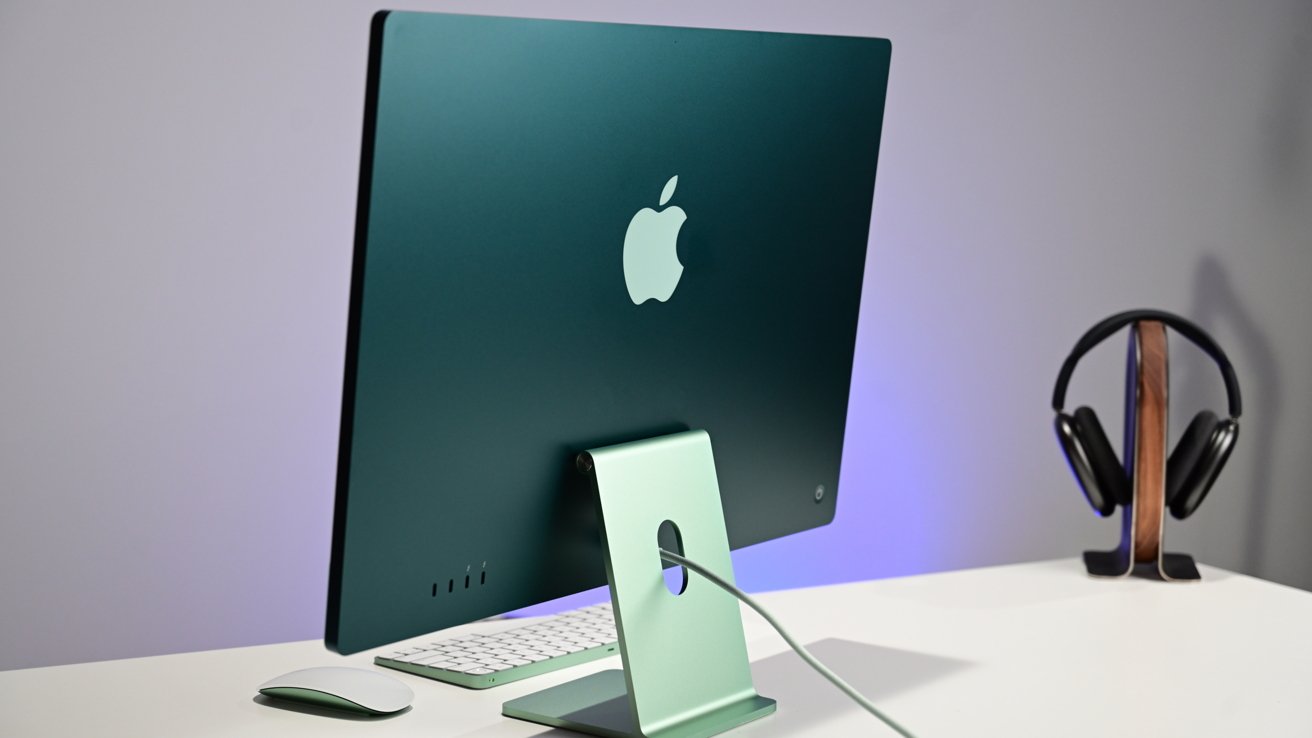 Зеленый компьютер Apple на столе с клавиатурой, мышью и наушниками на подставке.