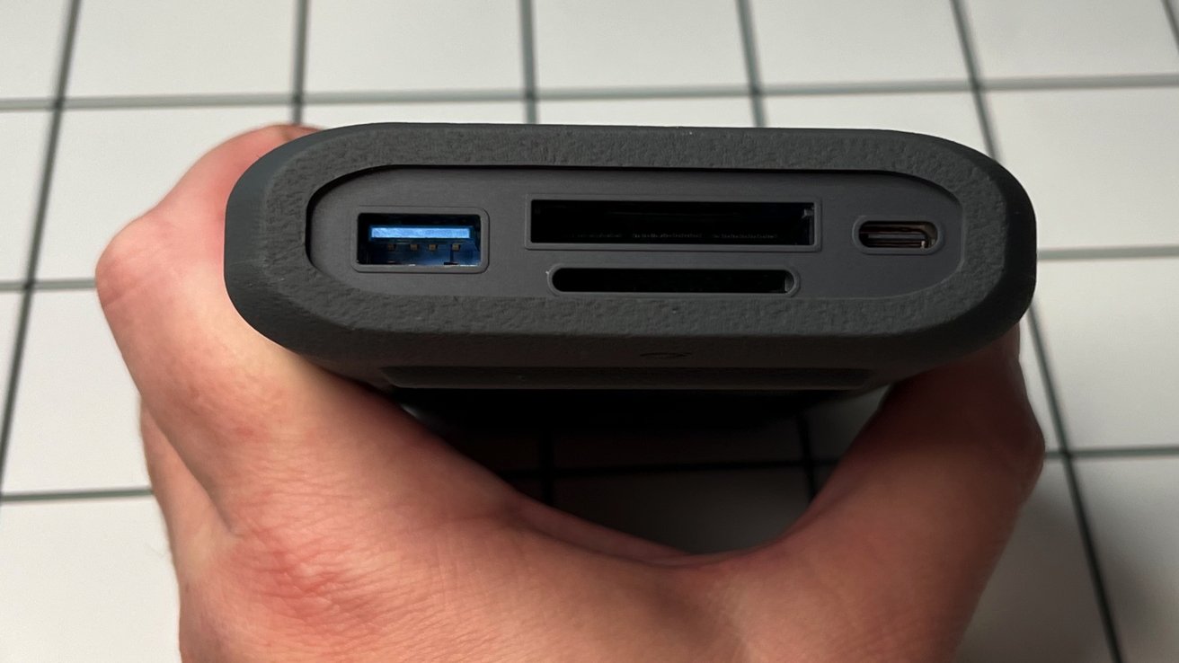 Рука держит черное устройство с портами, включая USB, SD-карту и USB-C, на мозаичном фоне.