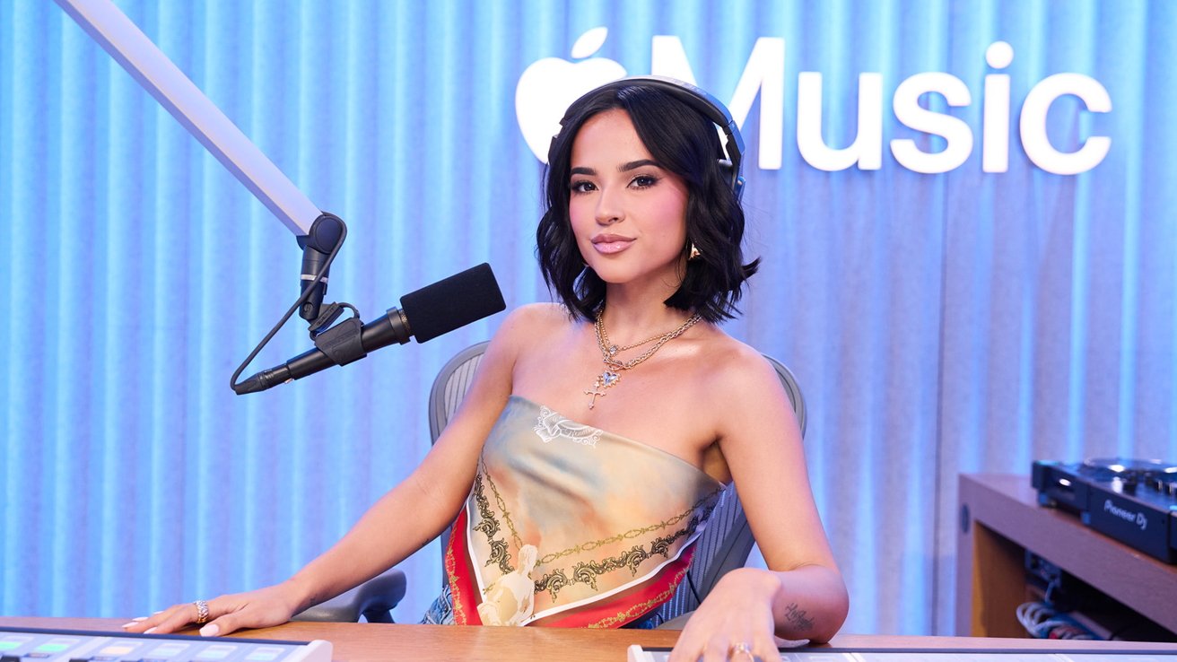 Человек сидит на радиостанции с наушниками, микрофоном и логотипом Apple Music на заднем плане, одетый в красочный топ без бретелек.