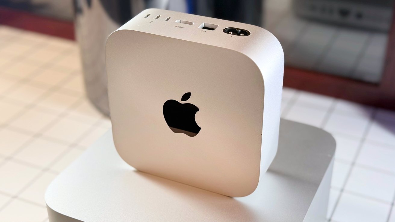 Устройство Apple в форме куба серебристого цвета с логотипом, закругленными углами и различными портами сверху, расположенное на отражающей поверхности.