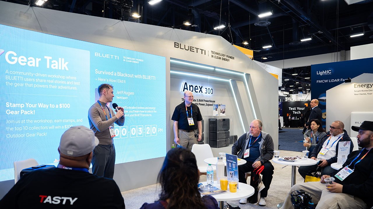 Представитель Bluetti с микрофоном обращается к сидящей аудитории в конференц-стенде CES 2025, на котором видны большие дисплеи и рекламные материалы.