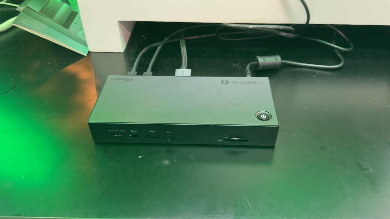 Черная док-станция UGREEN Thunderbolt с несколькими портами, включая USB, слоты для SD-карт и кнопку питания, расположена на отражающей поверхности зеленого оттенка.