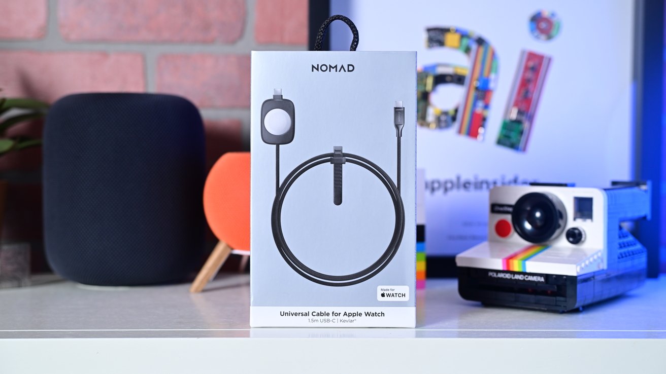 Коробка с универсальным кабелем Nomad для Apple Watch в окружении динамика, оранжевого устройства и винтажной камеры Polaroid на полке.