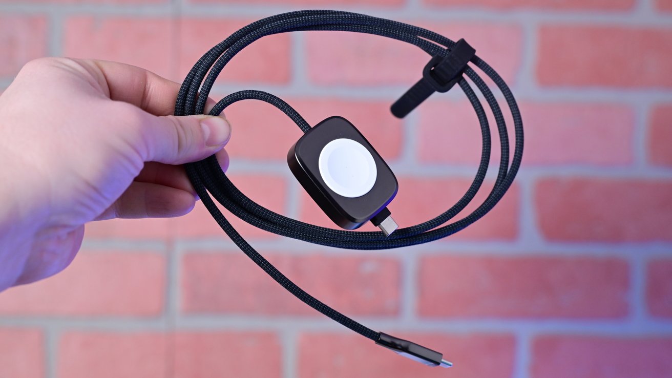 Рука держит черный зарядный кабель с разъемом USB-C и магнитной зарядной площадкой на фоне размытого кирпичного фона.