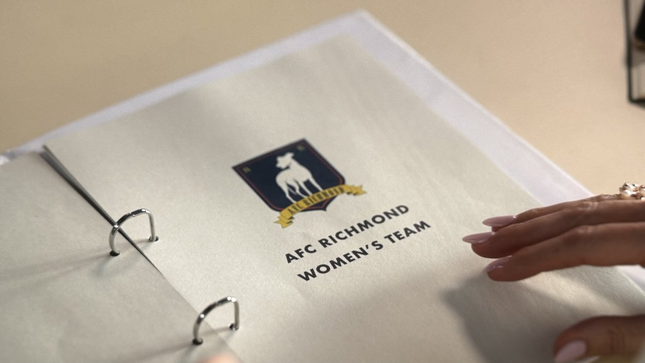 Папка с обложкой с надписью AFC Richmond Women's Team, на которой изображен логотип со стилизованной борзой. К странице прикасается рука человека с наманикюренными ногтями.