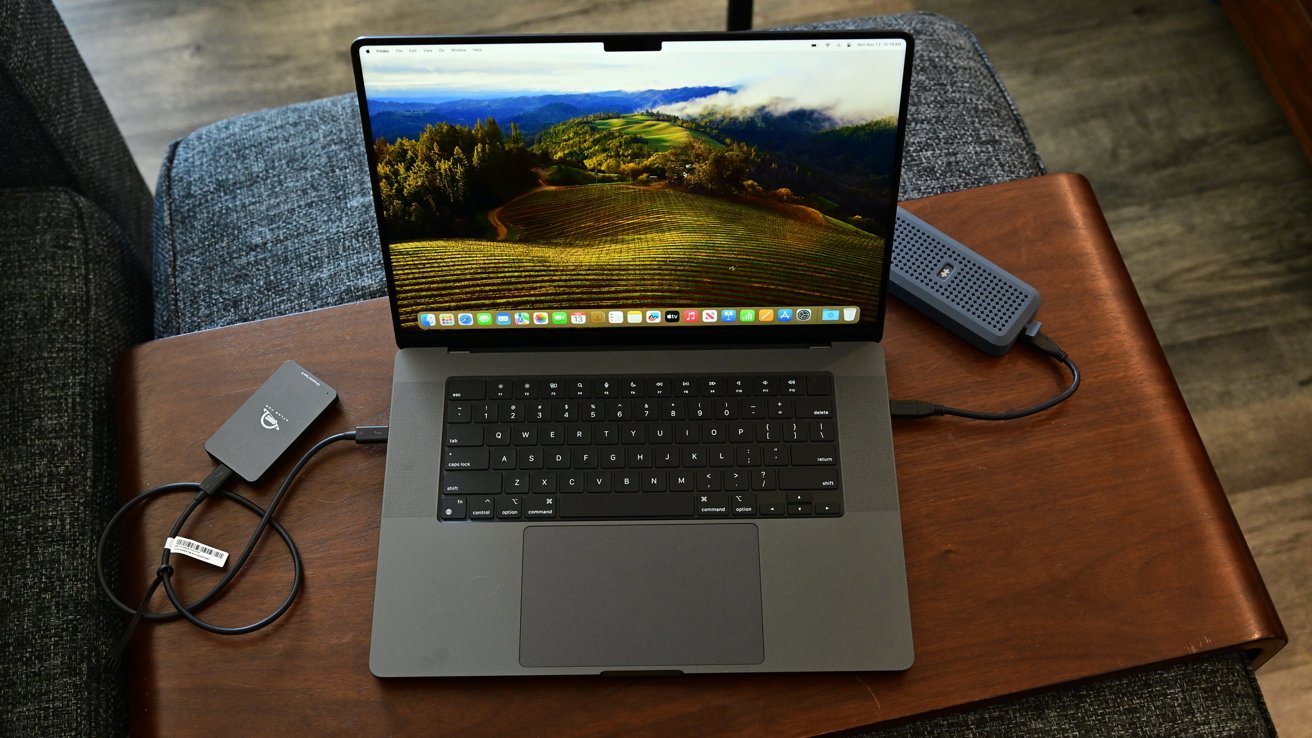 MacBook Pro на деревянном столе с пейзажными обоями, с двумя подключенными внешними накопителями, в окружении серой тканевой мебели.