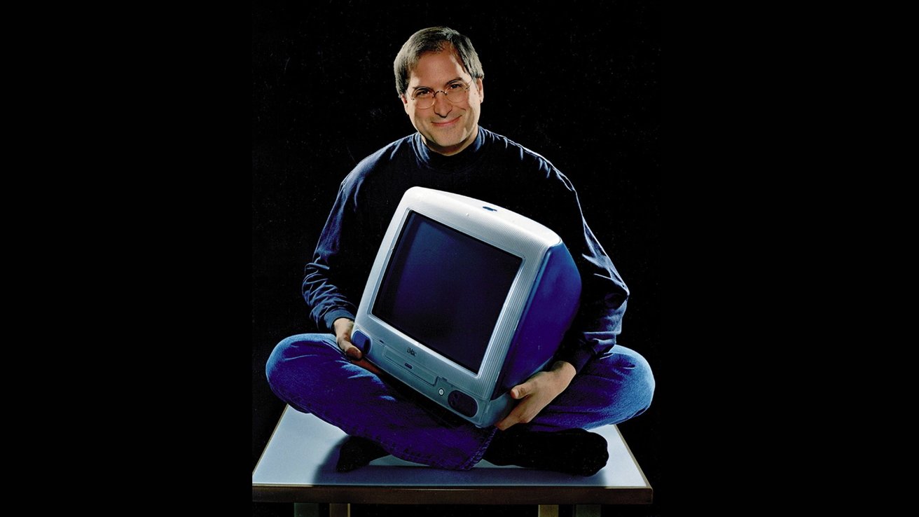 Улыбающийся Стив Джобс держит в руках оригинальный iMac, сидя со скрещенными ногами на платформе на темном фоне.